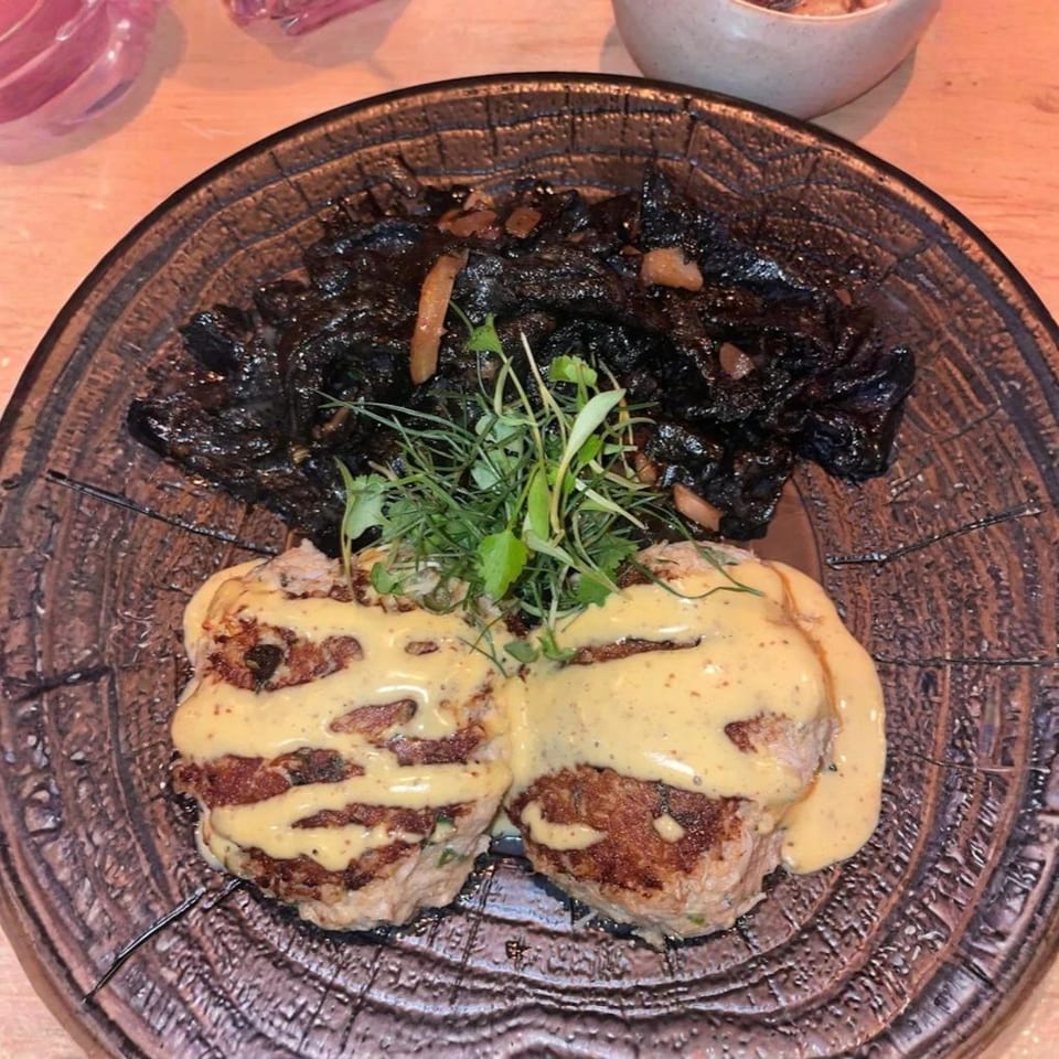Two crab cakes with yellow sauce and microgreens, alongside black squid ink pasta on a dark plate.